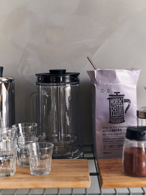 A coffee maker on a chopping board on an OMAR wire shelf, surrounded by POKAL glasses and a bag of coffee.