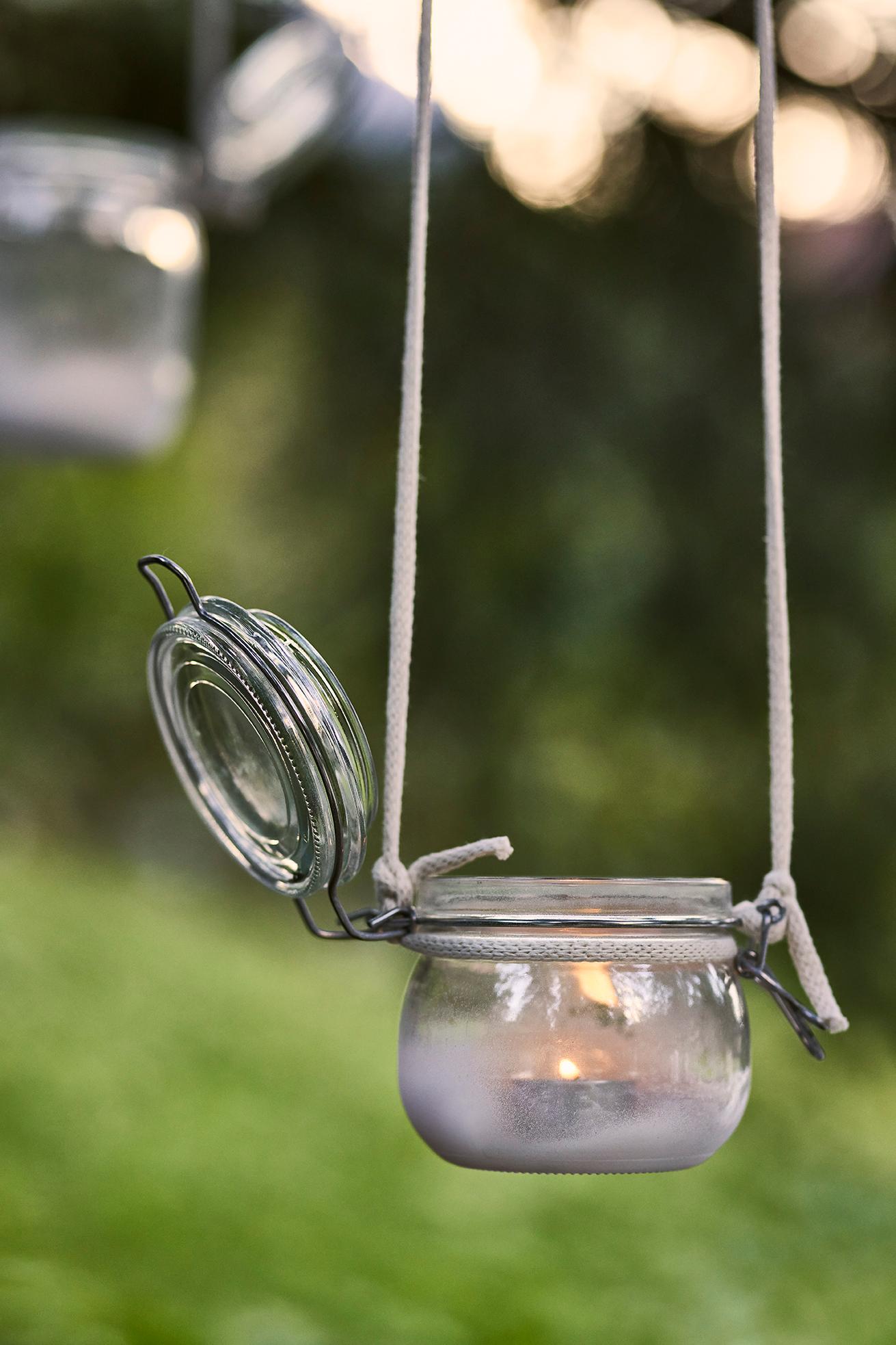 Nahaufnahme eines hängenden Glasbehälters mit Kerze als stimmungsvolle Outdoor-Dekoration vor grünem Hintergrund.