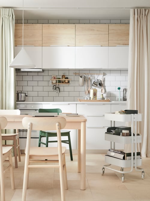 A table and coloured chairs beside a RÅSKOG trolley in front of a curtain-framed kitchen fitted with white VEDDINGE doors.