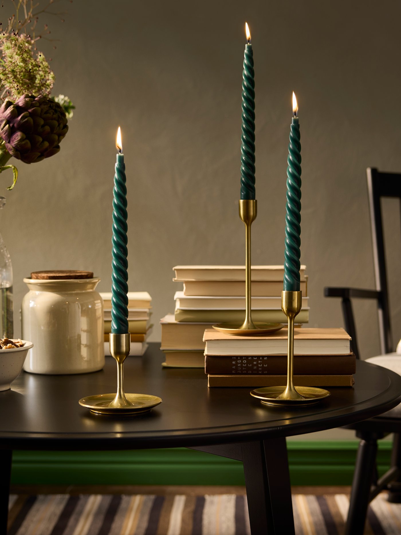 A set of 3 FULLTALIG candlesticks with green candles is placed on a black table beside a jar and a vase with flowers.