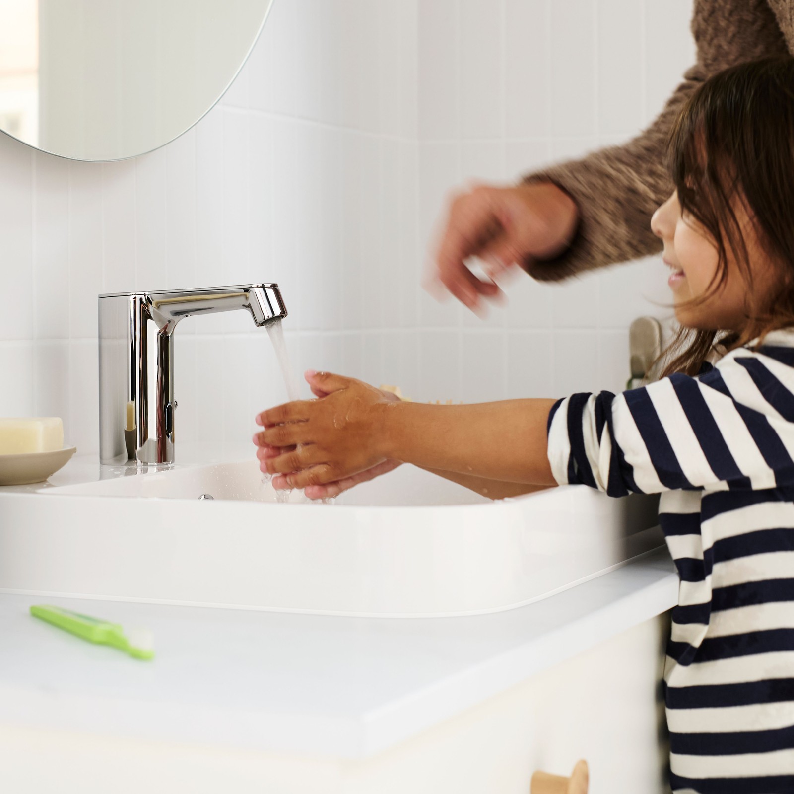 A child washing hands at a sink with an adult’s guidance, soap dispenser, toothbrush holder, and a bar of soap nearby.