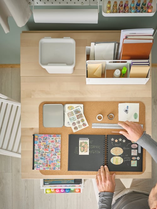 Workspace with wooden gateleg table, white desk organiser, fluorescent glitter paint, desk pad, bin with lid, chairs, person.