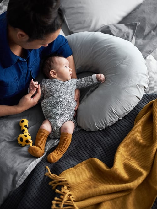 A person holding a baby resting on a grey LEN nursing pillow, with a yellow KLAPPA rattle and yellow OMTÄNKSAM throw nearby.