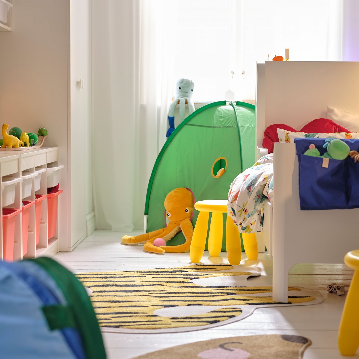 En BUSUNGE växasäng på ena sidan av ett litet sovrum, med en gul MAMMUT pall vid sidan av sängen och ett grönt lektält bakom den.