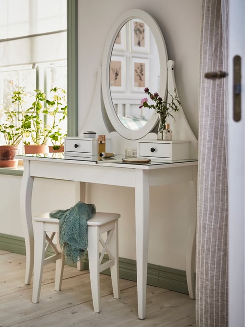 A white HEMNES dressing table with mirror has perfumes, flowers and makeup on top and a white stool underneath.
