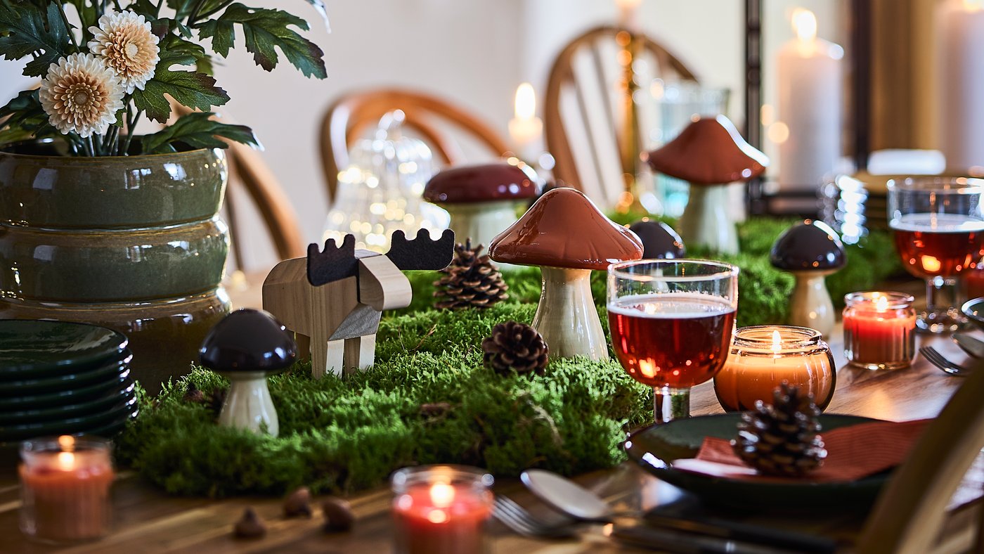 A woodland-themed dining table is set with HÖSTAGILLE decorative mushrooms and moose, lit candles, moss and pinecones.