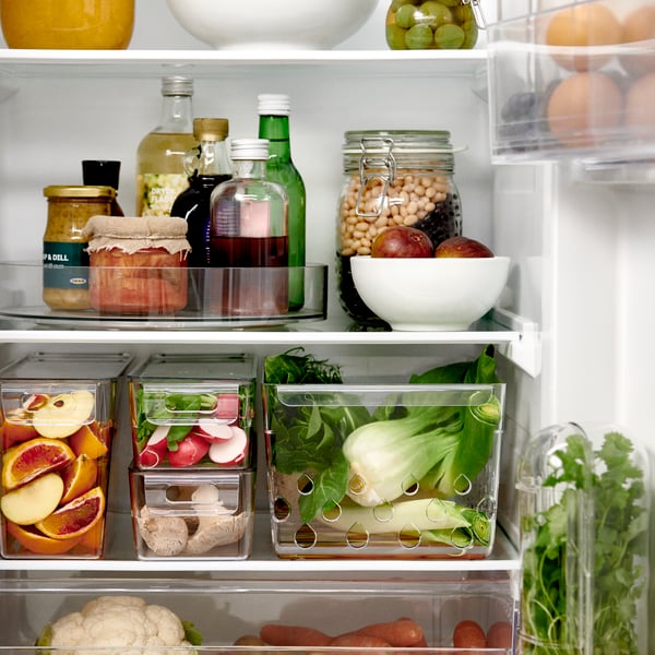 A transparent KLIPPKAKTUS storage basket holds fresh produce, surrounded by various bottles and jars in a fridge.