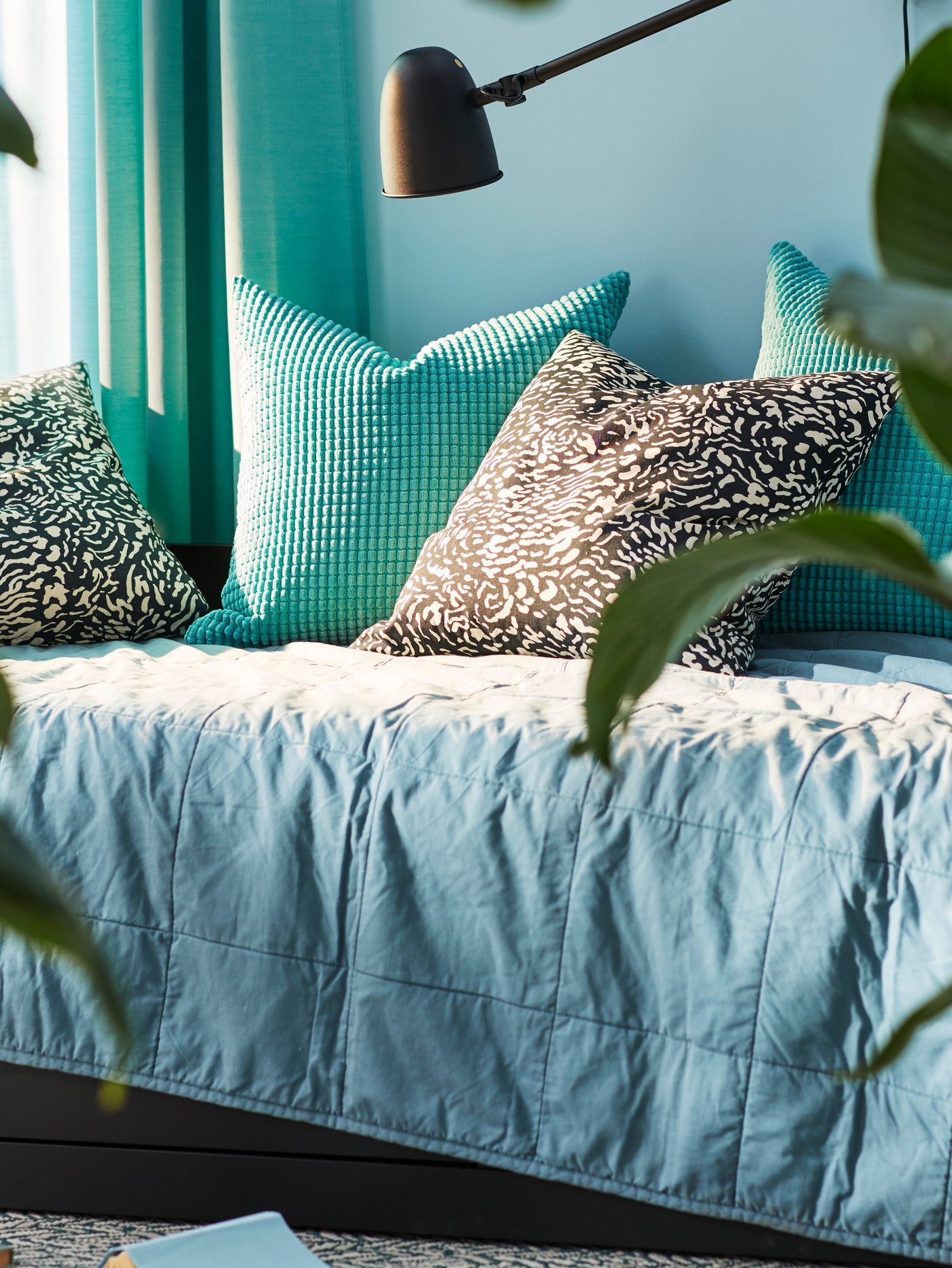Different cushions lie on a day-bed which is covered with a green GULVED bedspread. A SKURUP lamp is on the wall nearby.