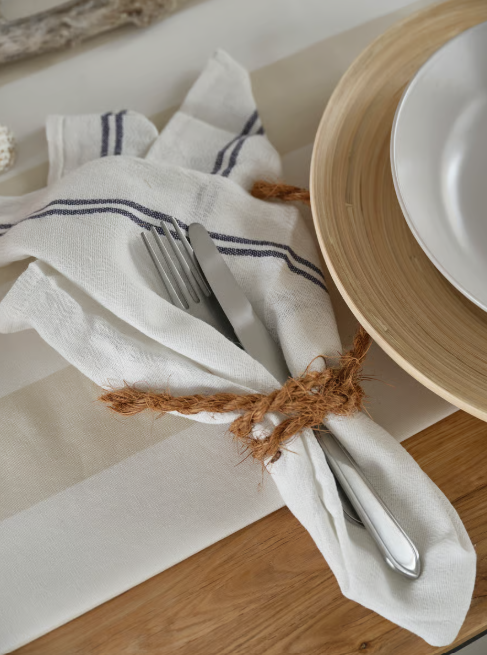 Gefaltete Stoffserviette mit Besteck, zusammengebunden mit Kordel, neben weissem Teller auf hellem Holztisch.
