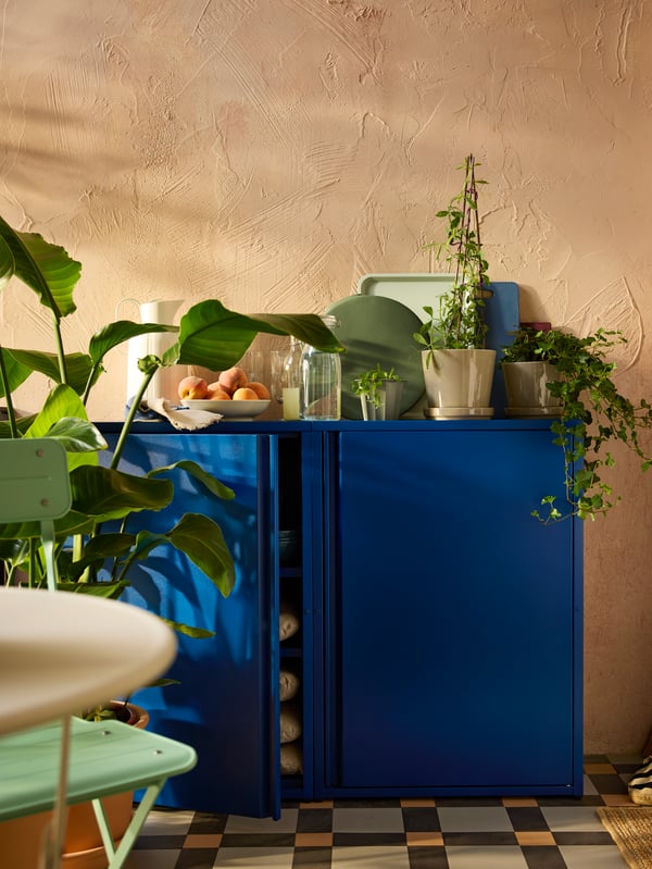 A balcony wall with two dark blue SUNDSÖ cabinets with plants, glasses, and an off-white TAGGÖGA vacuum flask on top.