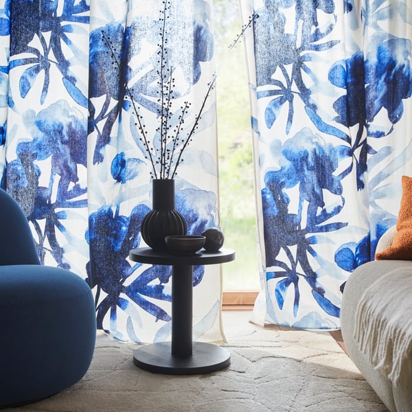 Dzīvojamā istaba ar baltiem/ziliem SKOGSNÄSSLA aizkariem ar ziedu rakstu, kas karājas blakus zilam atzveltnes krēslam un galdam.
