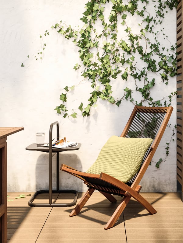 A wooden lounger with black mesh and a green cushion and a side table with a magazine, sunglasses and a glass of water.