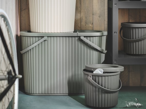 The image shows a section of an interior space, featuring a ribbed metal laundry basket with a curved top edge and a single gray handle on the side. The basket is placed on a teal-colored carpeted floor against a wall with vertical wooden paneling partially obscured by what appears to be the edge of a white door or divider. To the left, there is the partial view of another object with intricate patterns, possibly part of furniture or home decor.