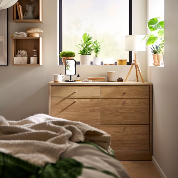 TONSTAD chest of six drawers in oak veneer in the corner of a bedroom below a window. Foot end of a bed in foreground.