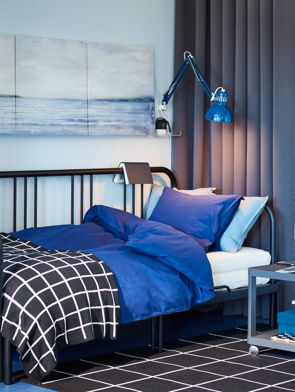 A black day-bed with blue bed textiles, block-out curtains, a black and white rug and a work lamp.