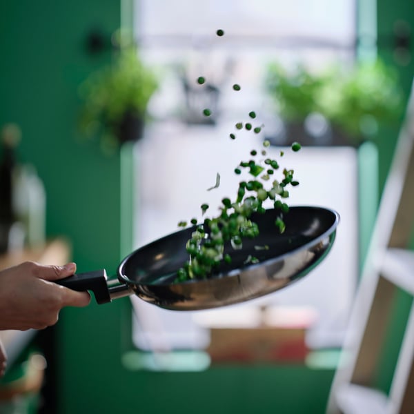 A hand is holding a stainless steel MIDDAGSMAT frying pan with a non-stick coating, tossing the veggies in it up in the air.