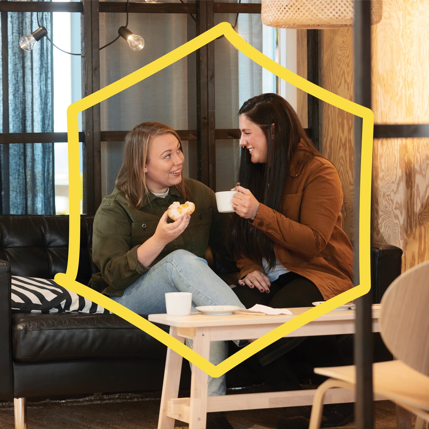 Two women having coffee on a black sofa in an IKEA restaurant. A yellow frame in the shape of a hexagon on top of the image.