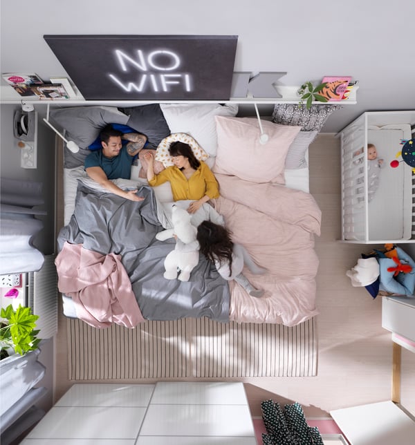 A couple lie on a large bed where their daughter is playing with an animal soft toy. A baby lies in a SUNDVIK cot nearby.