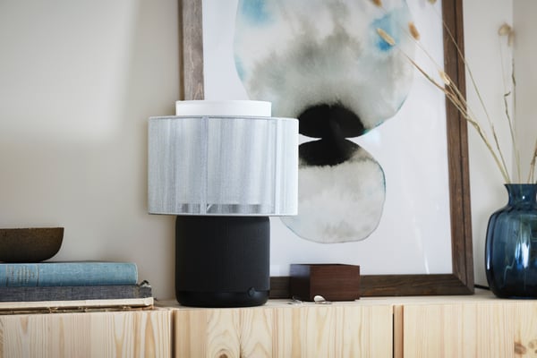 A SYMFONISK table lamp with WiFi speaker in white/gray placed on a bedside table.