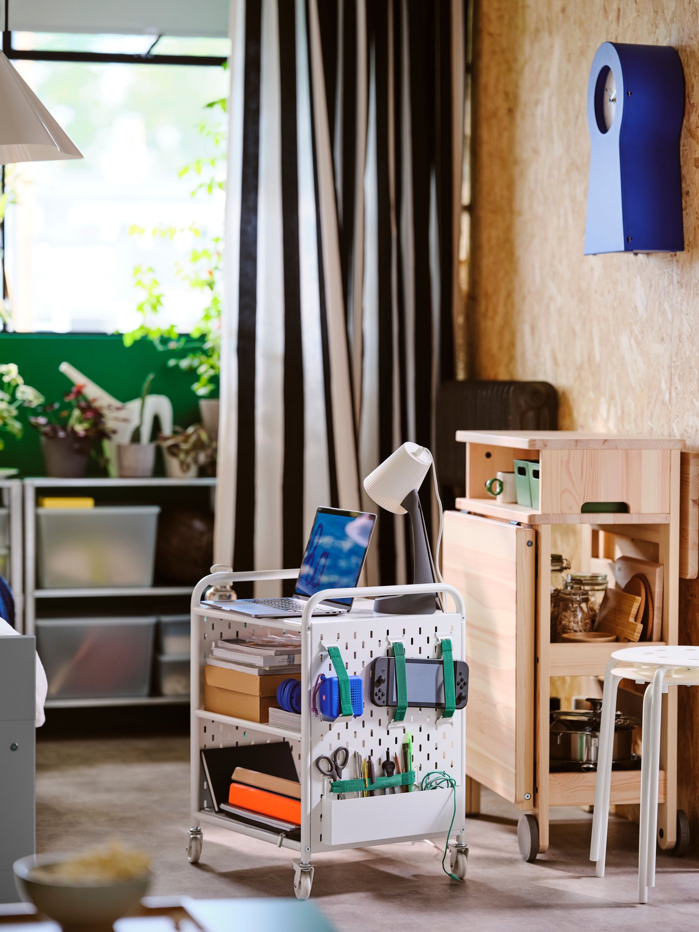 Pracovní prostor s pojízdným bílým vozíkem plným kancelářských potřeb a notebookem, vedle dřevěného úložného stolku, v pozadí rostliny a police.