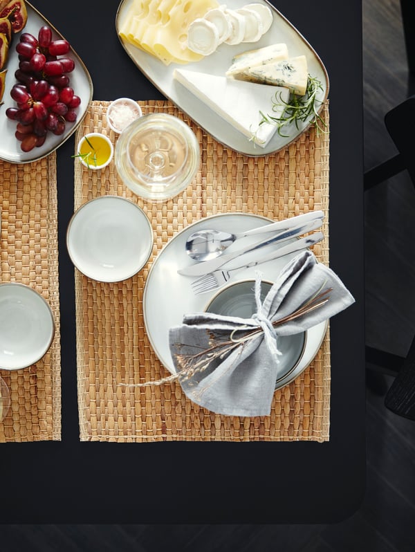 A table that’s set with place mats made from water hyacinth, grey dinnerware and two serving plates with cheese and grapes.