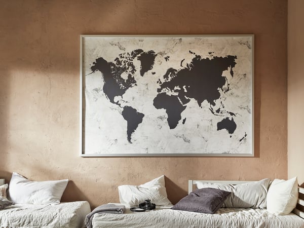 A large, framed, borderless world map in black and white, hangs on a beige wall above two day beds.