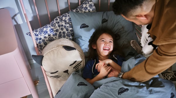 A girl gets cuddled in bed by her father, and smiles widely at him as she lays comfortably in a BARNDRÖM duvet.