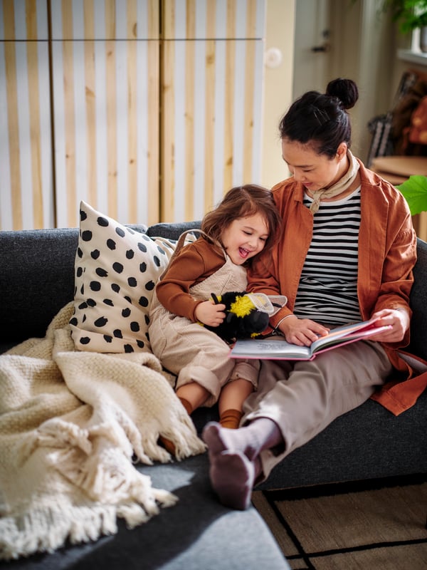 A person reads a story to a small child who is leaning against a cushion with an ODDNY cushion cover on a sofa.