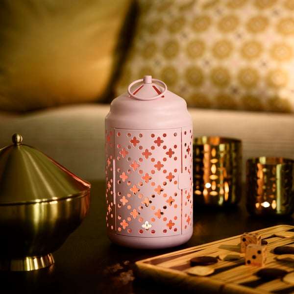 A pink GOKVÄLLÅ tealight lantern in powder coated steel, with a lit candle, next to a gold-coloured GOKVÄLLÅ serving bowl.