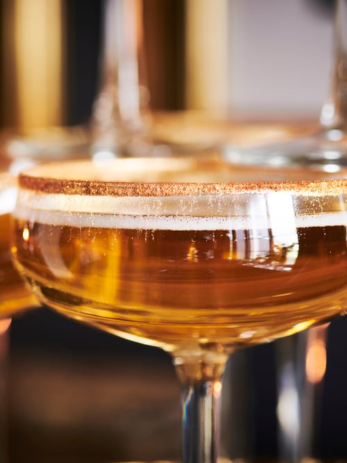 A close-up of STORHET champagne coupe shows it is filled with an amber-coloured liquid and has edible gold on its rim.