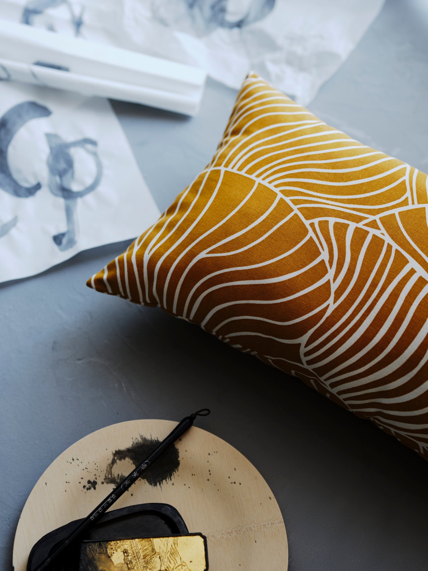 A dark yellow SVAMPMAL cushion with a white mushroom print is on a pale grey wooden surface with a paintbrush and paper.