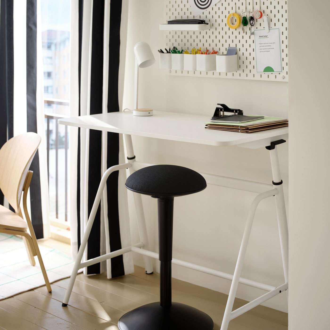 A white GLADHÖJDEN sit/stand desk is in a raised position, by a white wall with pegboard storage in a student room rental.