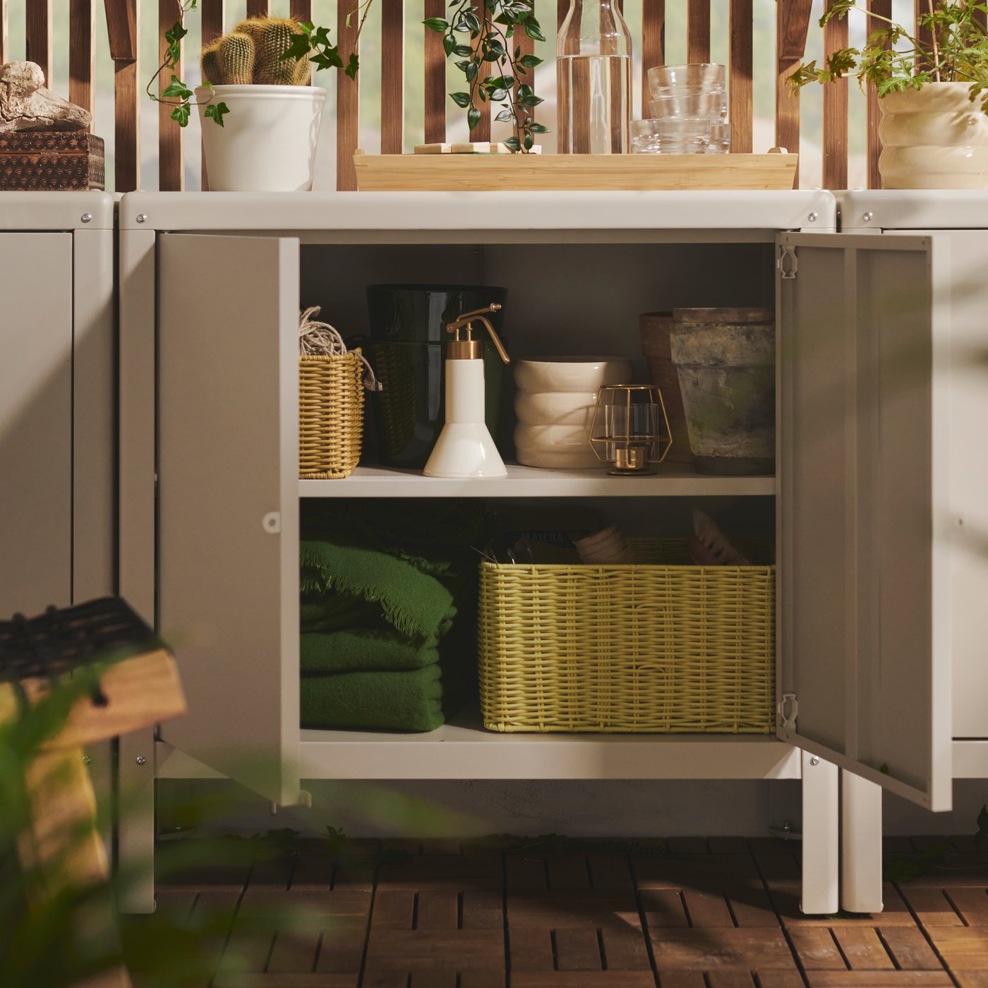 A balcony with a beige KOLBJÖRN cabinet displaying blankets, gardening tools and storage baskets in its open shelves.