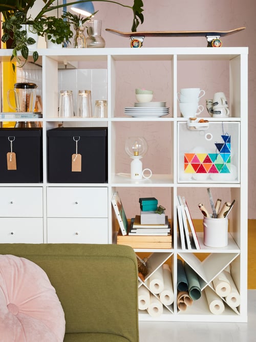 Living room with shelving unit, decorative objects, personal items, a sofa, a cushion, dinnerware, plants, boxes, lamps.