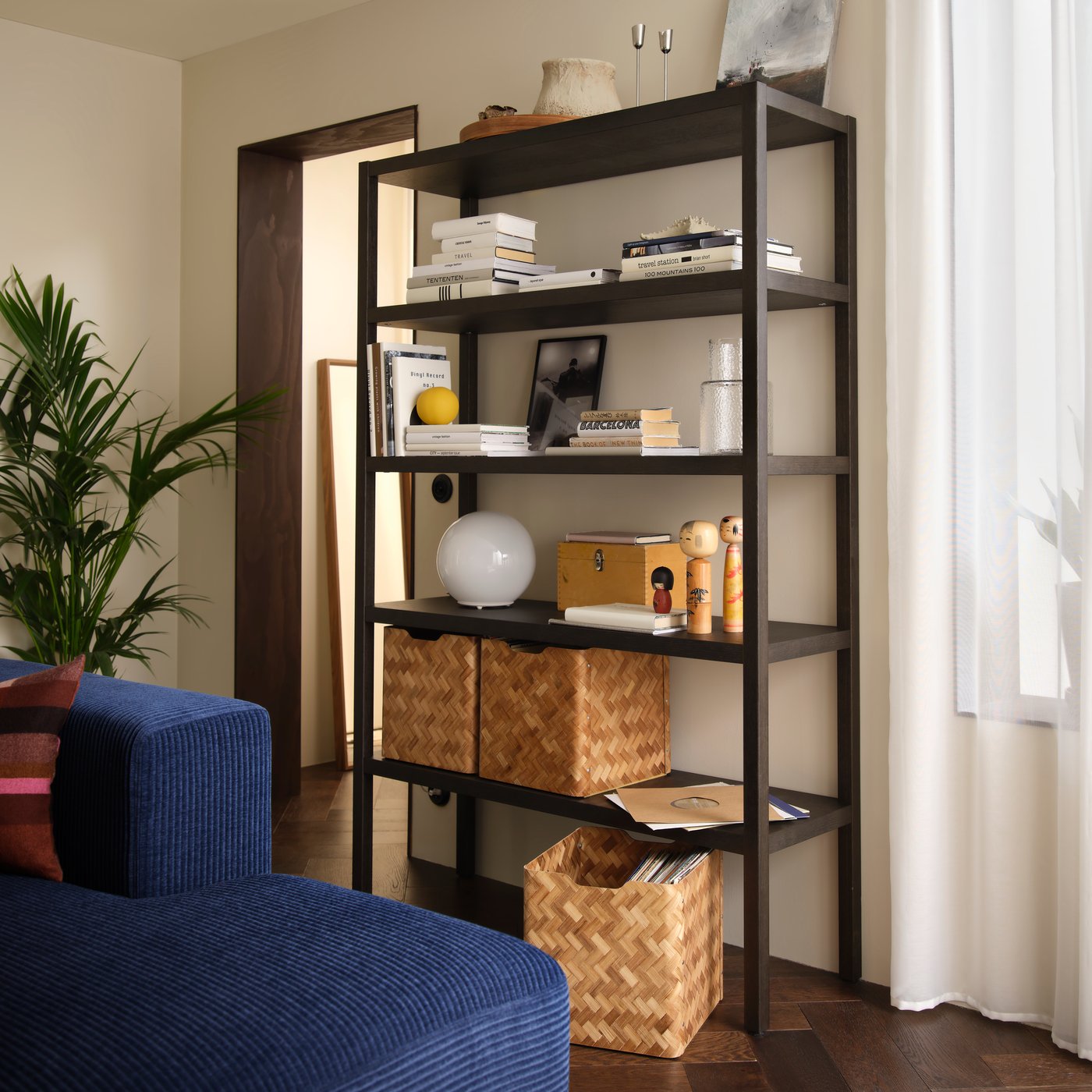 A living room with a dark-brown oak-veneer STOCKHOLM 2025 shelving unit. It is filled with decor, books, and baskets.