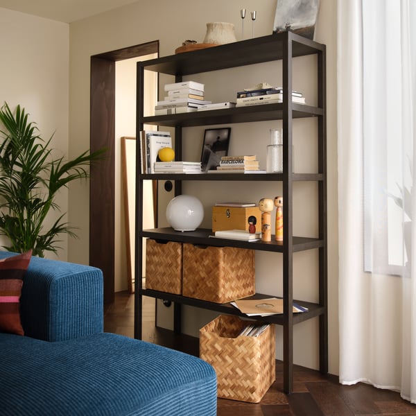 A living room with a dark-brown oak-veneer STOCKHOLM 2025 shelving unit. It is filled with decor, books, and baskets.