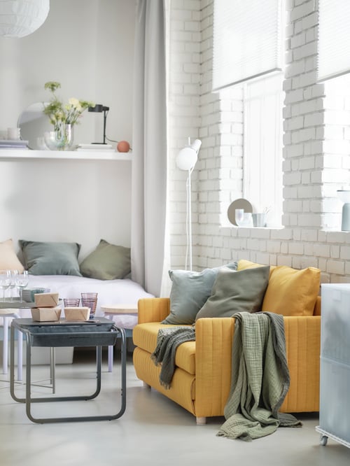 Wohnraum mit einem FRIDHULT Bettsofa mit Kissen und einem Plaid, u. a. mit einer HÅRSLINGA Standleuchte mit Lampe und einem OLSERÖD Beistelltisch.