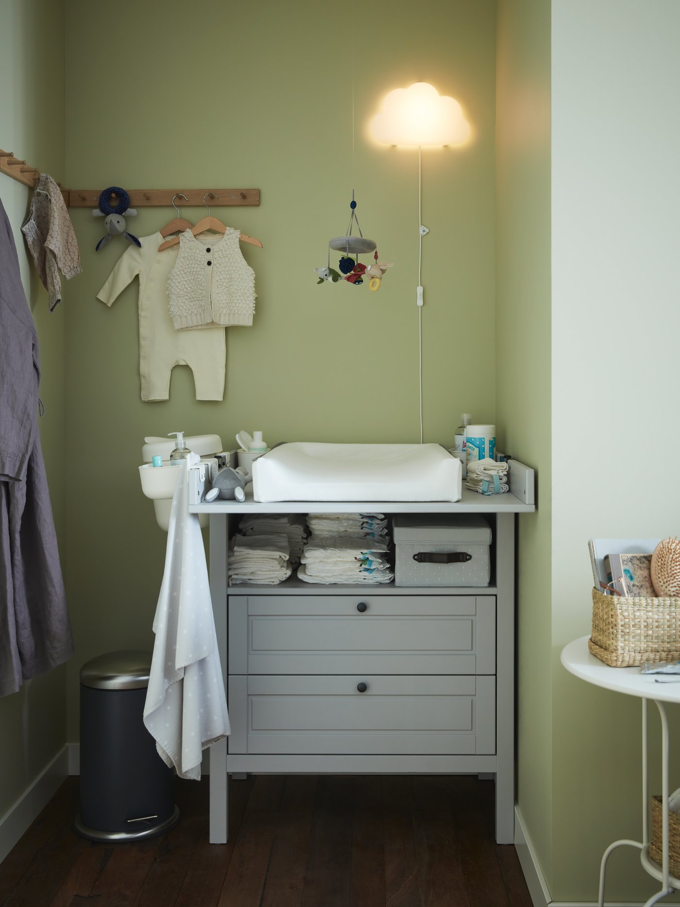 A grey changing table/chest of drawers, oak racks with baby clothes hanging, a babycare mat, a cloud white LED wall lamp.