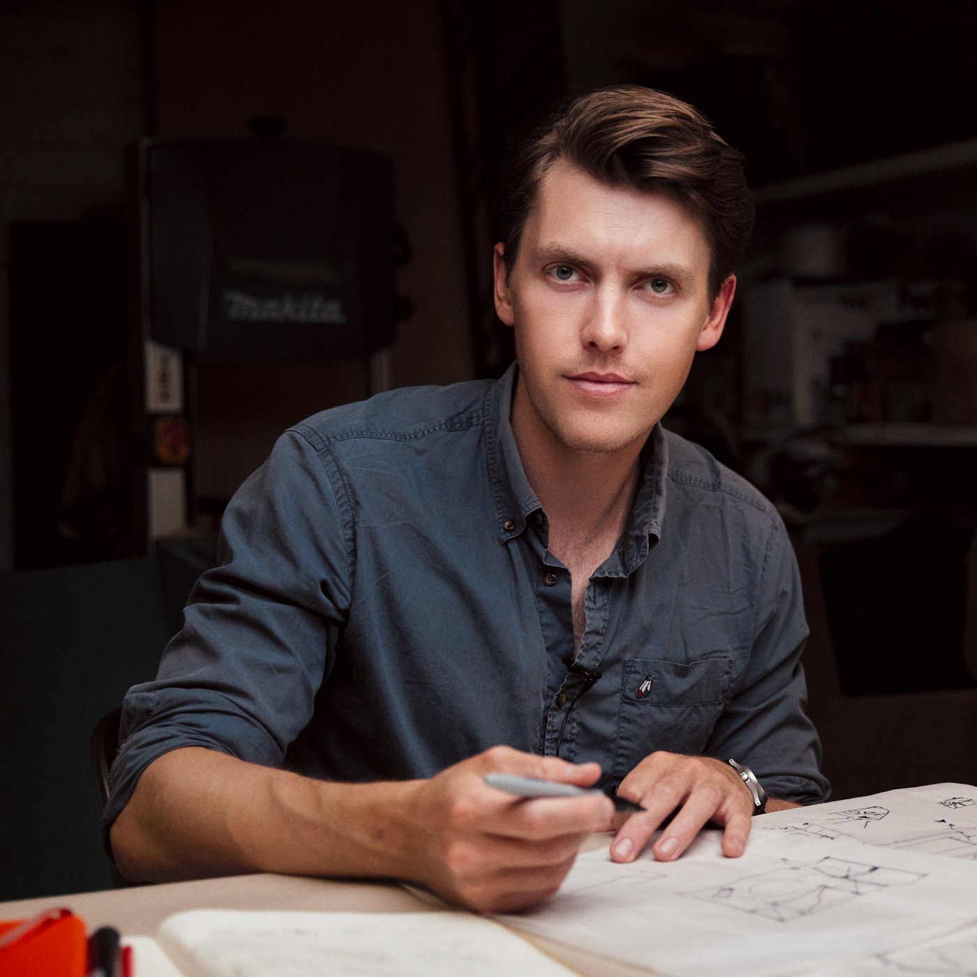 A man sitting behing a desk with a pen in his hand. He is looking into the camera.