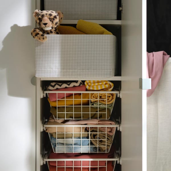 STUK storage case in a wardrobe, filled with folded clothes and a soft toy on top, beside hanging garments.