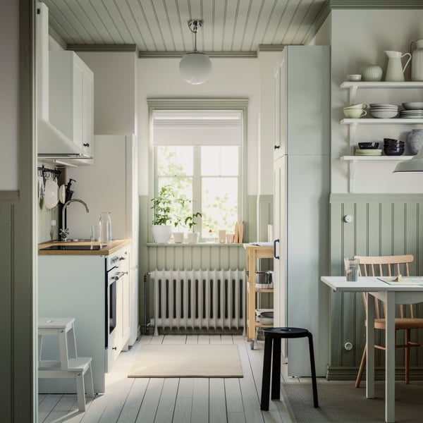 A small parallel kitchen with a window has white KNOXHULT cabinets, a dining table, a rug on the floor and a step stool.