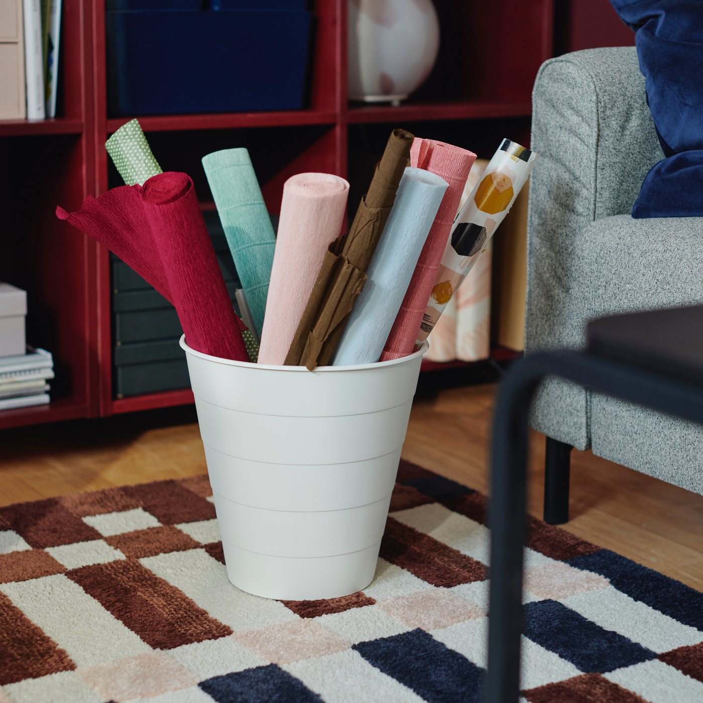 Poubelle FNISS blanche contenant des rouleaux de papier coloré, sur un tapis MARKBLÅMME et devant des étagères rouges.