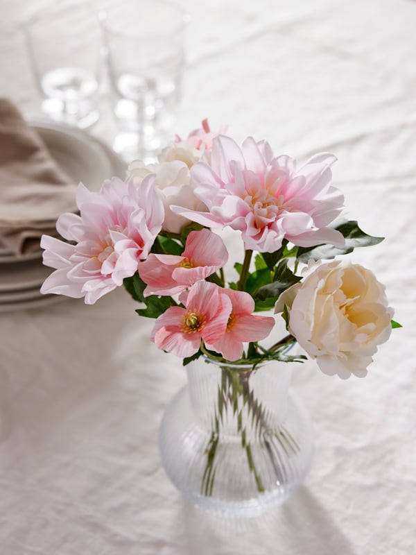 A pink poppy and white peony and pink dahlia SMYCKA artificial flowers stand in a clear glass PÅDRAG vase on a white surface.