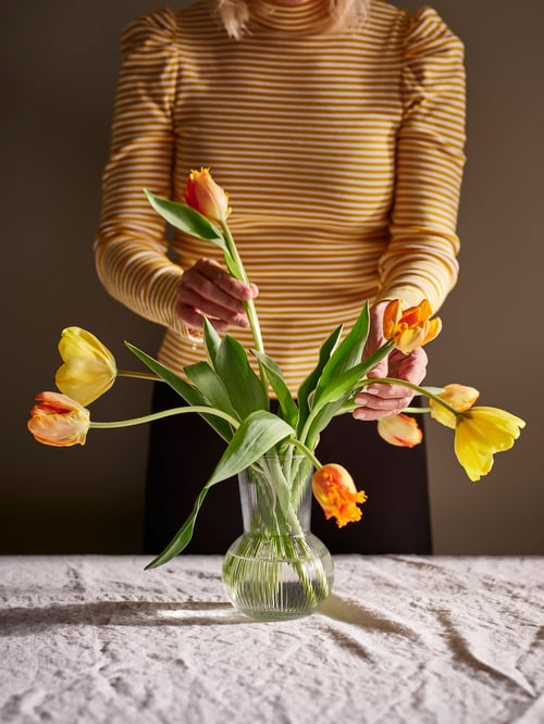 Person arranging tulips inside an elegant glass vase with round base and vertical ridges, on a table with white tablecloth.