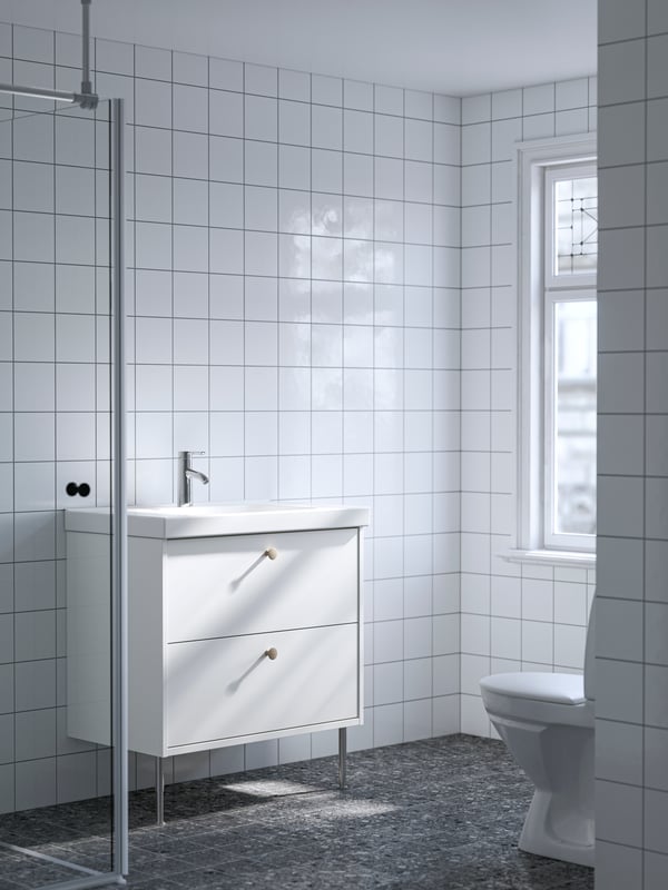 A white HAVBÄCK wash-stand with two drawers and an ORRSJÖN wash-basin is placed in a white-tiled bathroom with a window.