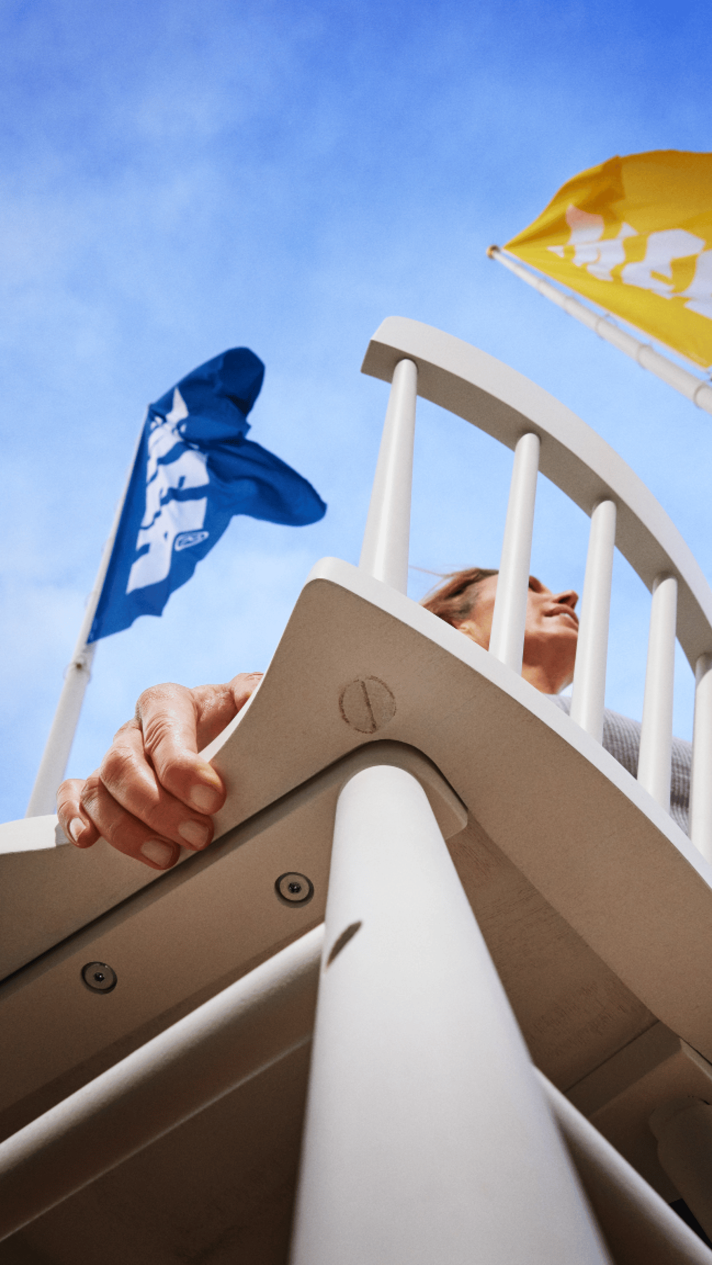 Una persona tiene le mani sui braccioli di una sedia, circondata da bandierine IKEA colorate che sventolano contro un cielo azzurro.