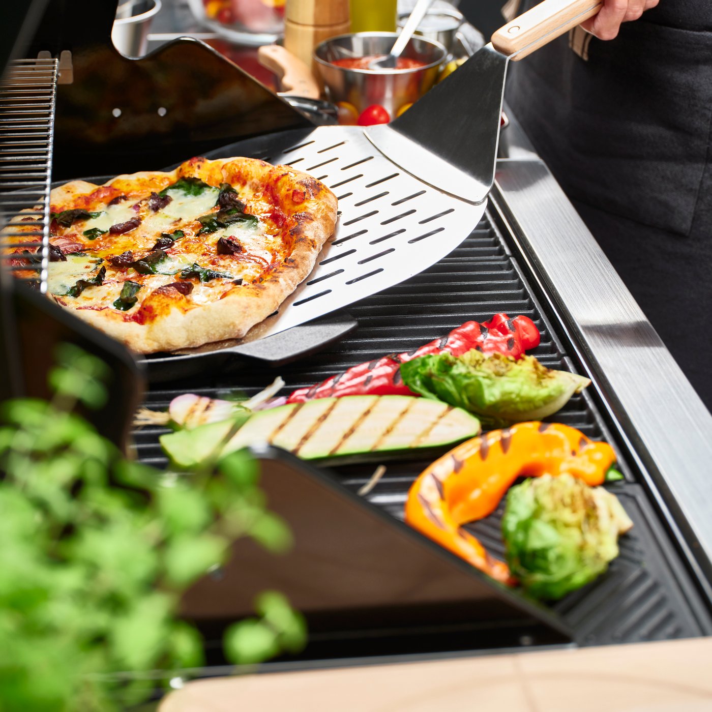 Pizza on a grill lifted with a spatula, surrounded by grilled vegetables and herbs nearby.