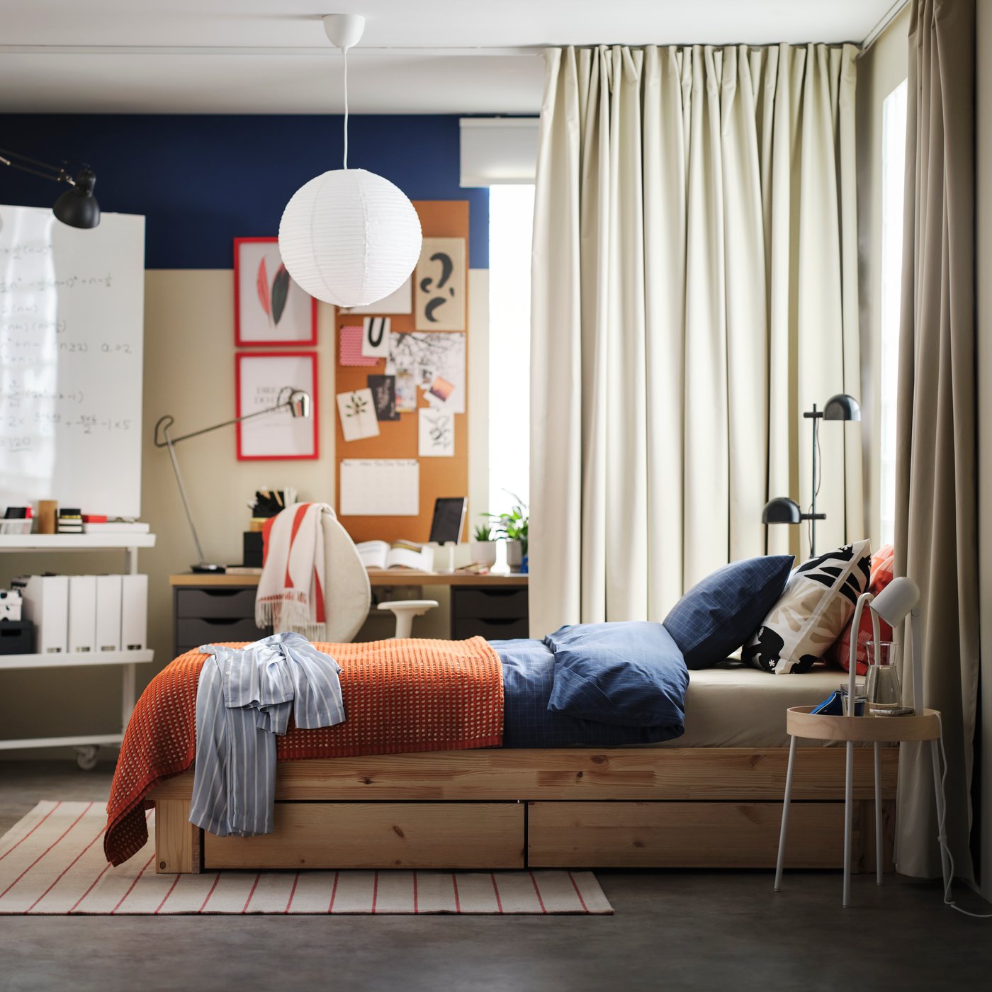 A pine GLAMBERGET extendable bed with storage in a bright bedroom with a BENGTA block out curtain between it and a desk area.