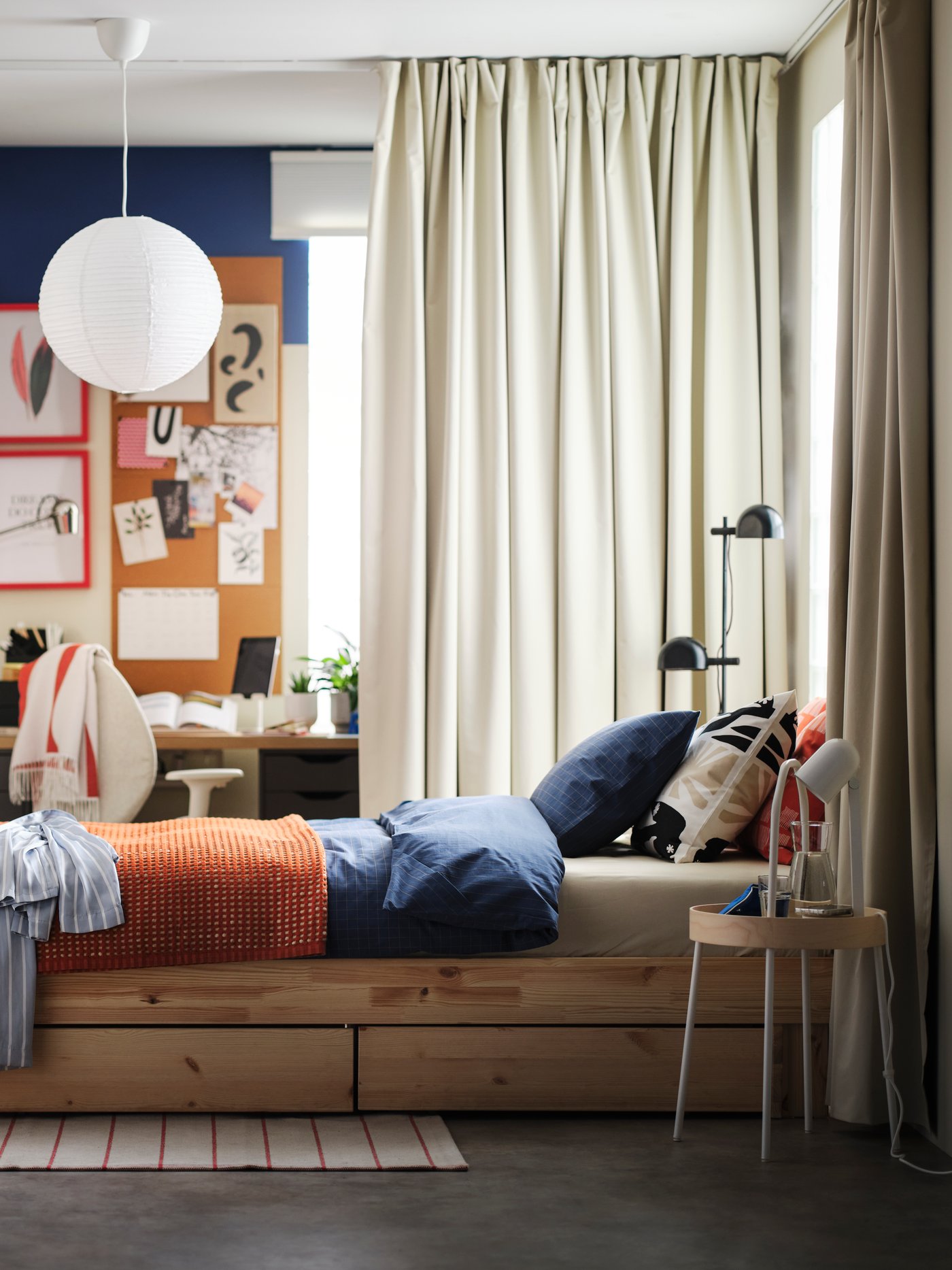 A pine GLAMBERGET extendable bed with storage in a bright bedroom with a BENGTA block out curtain between it and a desk area.