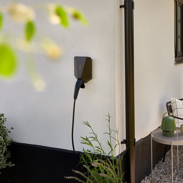 Ein Ladegerät für Elektrofahrzeuge (EV), das an der Aussenseite eines Hauses angebracht ist. Ein Zweig mit grünen Blättern ist links oben im Bild unscharf.